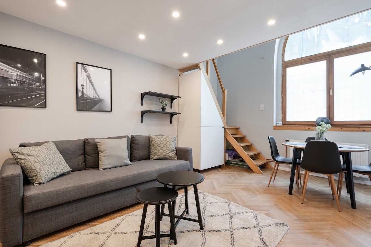 A modern living area features a gray sofa adorned with decorative cushions. Two small black tables are positioned centrally, and minimalistic shelving displays framed artwork. Natural light enters through large windows, illuminating the hardwood floor and staircase leading to an upper level.