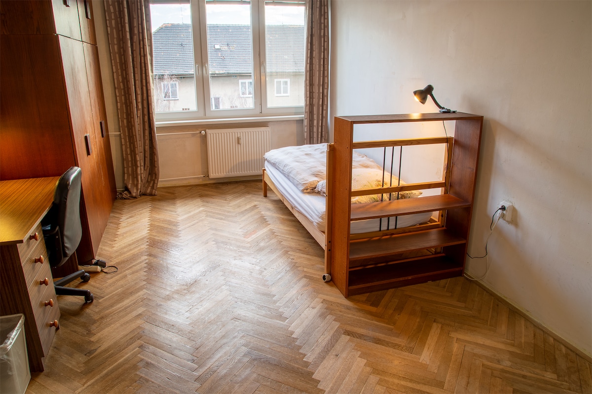 The room features a spacious bed with a wooden frame and a built-in bookshelf positioned next to it. A desk with a chair is visible against one wall, while large windows allow natural light to illuminate the wooden floor. Neutral-toned curtains frame the windows.