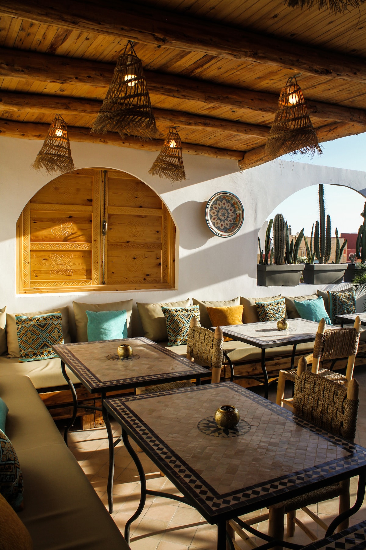 A charming seating area features natural wood ceiling beams and decorative woven light fixtures. Comfortable cushioned seating surrounds intricate patterned tables. Potted cacti add a touch of greenery, while wooden shutters create a warm, inviting space filled with natural light.