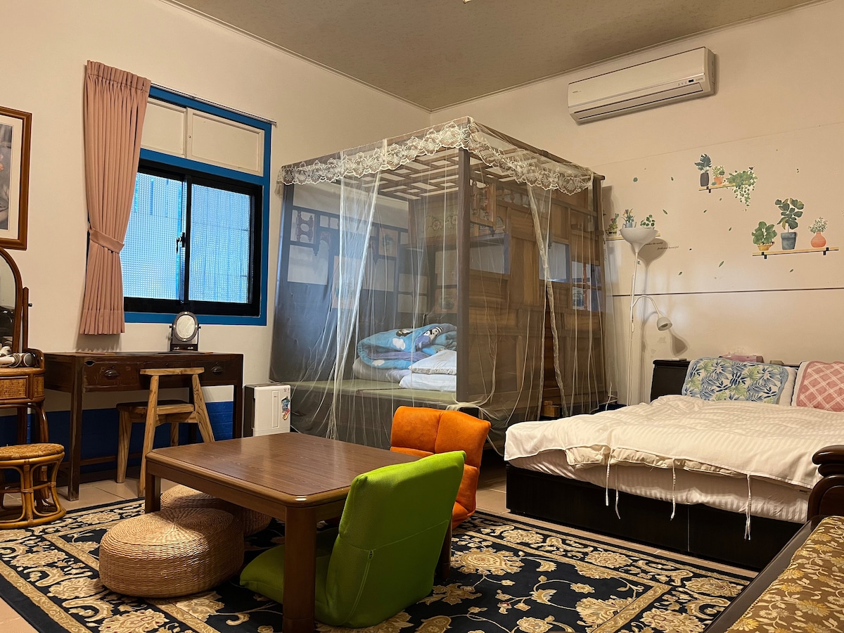 A stylish room features a spacious bed accompanied by a cozy sofa. A wooden table sits in the center, surrounded by colorful cushions. A bookshelf is enclosed in a protective netting, with a window allowing natural light to fill the space.