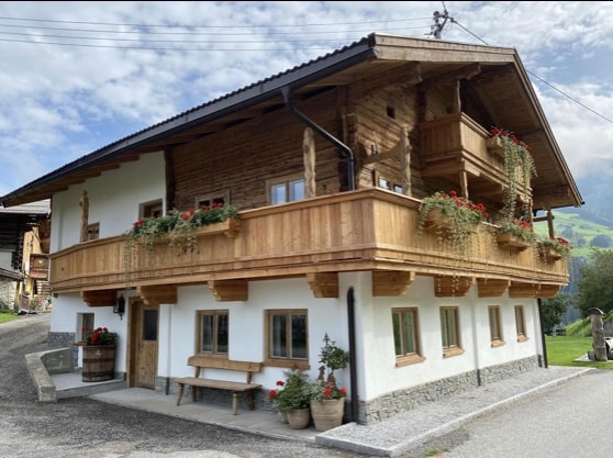 A charming wooden chalet is depicted, featuring natural wood siding and a spacious balcony adorned with flower boxes. The structure's ground floor is enhanced by large windows, allowing for natural light. A stone pathway leads up to the entrance, surrounded by scenic greenery.