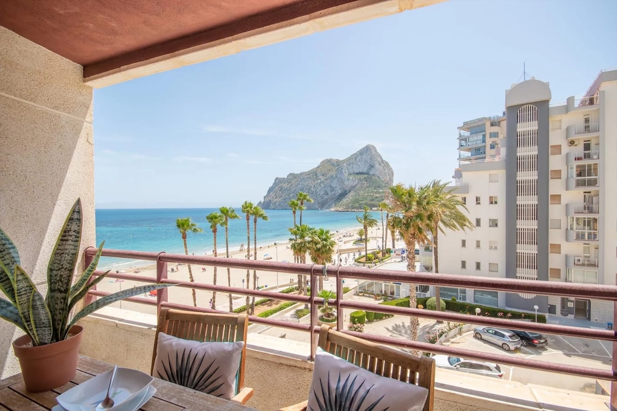 A balcony offers a breathtaking view of the beach and sea, framed by palm trees. The Peñón de Ifach stands prominently in the distance. Outdoor furniture enhances the space, inviting relaxation while enjoying the coastal scenery.