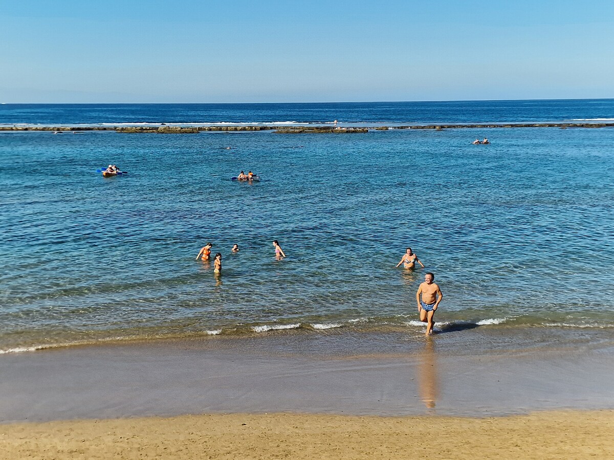 The image captures a sandy beach with people enjoying the clear water. Some individuals are swimming, while others wade in the shallow surf. The ocean extends to the horizon under a clear blue sky, creating a serene coastal scene.
