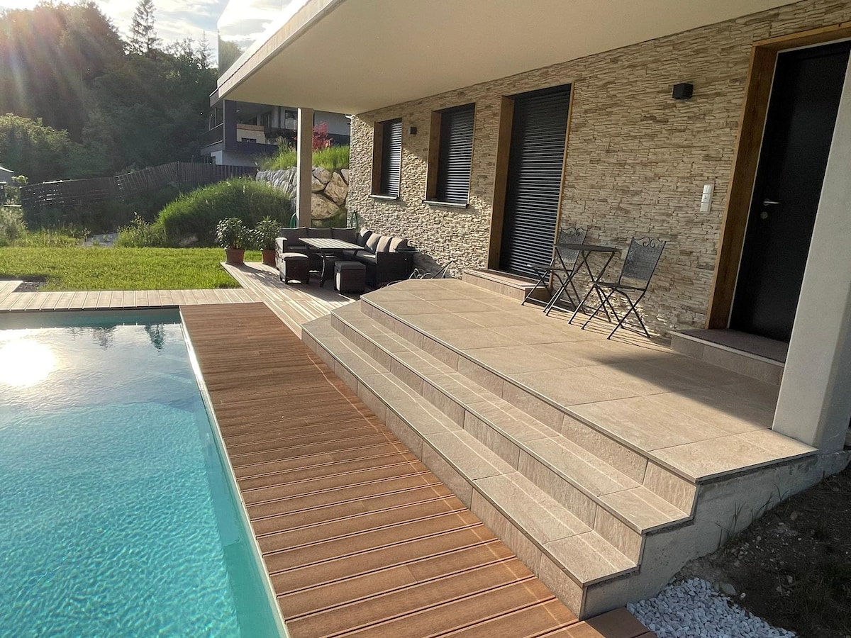 A modern outdoor area features a private pool adjacent to a wooden deck. A small seating arrangement with a couch and two chairs overlooks the pool, while steps lead to the entrance of the apartment. Lush greenery is present in the background.