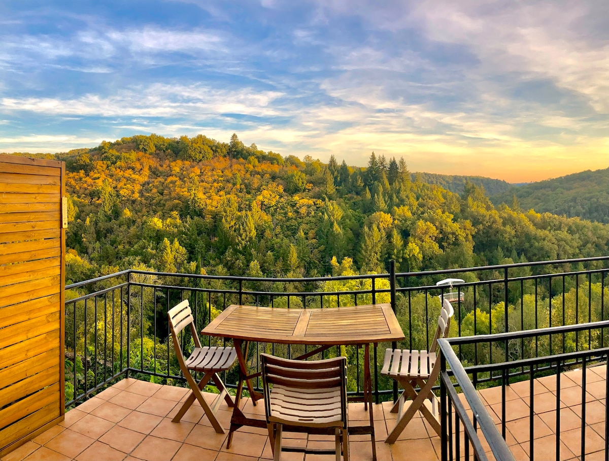 A terrace is visible, featuring a wooden table set with two chairs, surrounded by a scenic view of rolling hills covered in lush greenery. The sky displays warm hues of sunset, adding a serene backdrop to the natural landscape.