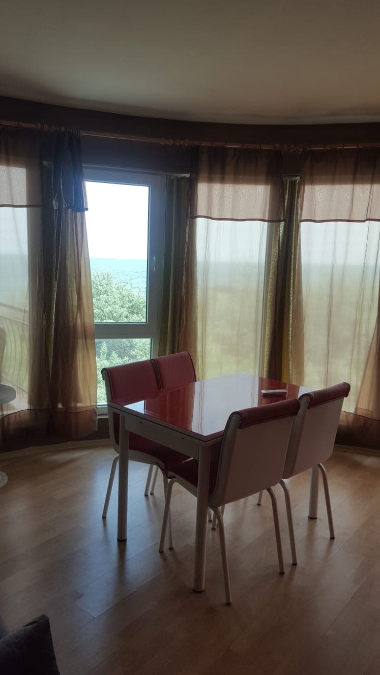 A dining area features a small table with four chairs, positioned near a large window draped with sheer curtains. The view beyond the window reveals greenery and a distant glimpse of the sea, allowing natural light to brighten the space.