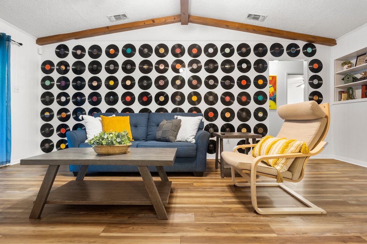 A spacious living area is highlighted by a feature wall adorned with an array of vinyl records. A comfortable blue sofa faces a wooden coffee table, while a light-colored chair with a striped cushion provides additional seating. Natural light flows in from nearby windows.
