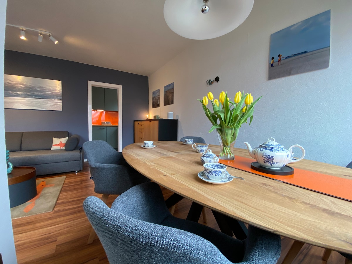 A dining area features a round wooden table surrounded by upholstered chairs. A vase filled with fresh tulips sits prominently on the table, accompanied by a tea set. Artwork depicting coastal scenes adorns the walls, while a gray sofa is visible in the background.