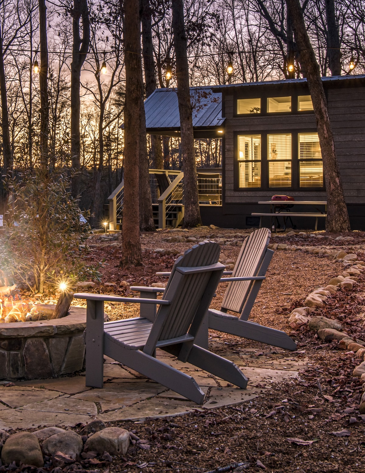 An outdoor seating area features two grey Adirondack chairs beside a circular stone fire pit. The setting is surrounded by trees, with warm lights strung overhead. The tiny home is visible in the background, highlighting its large windows that reflect the soft evening light.
