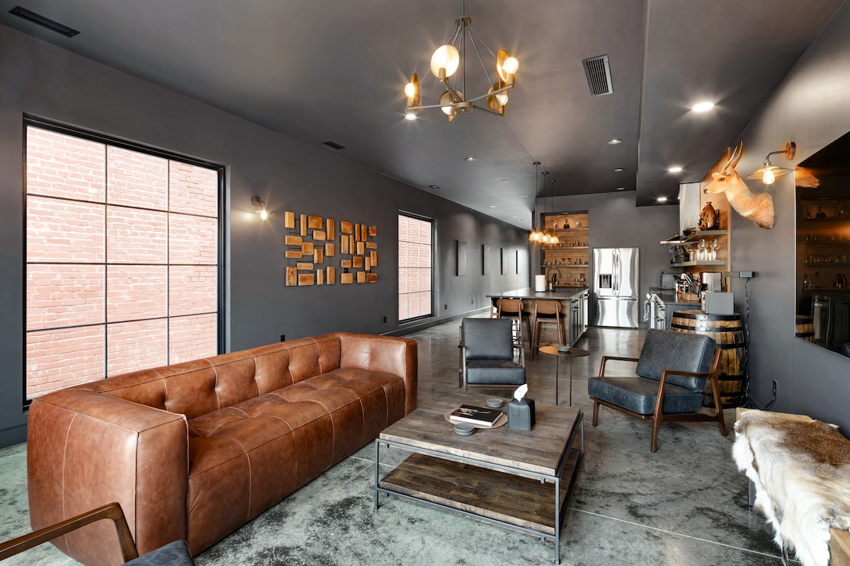 An open living and dining space is characterized by a rich brown leather sofa and wooden accents. Industrial-style lighting illuminates the room, while large windows allow natural light to flood in. A wooden dining table and a kitchen area featuring stainless steel appliances complete the inviting atmosphere.