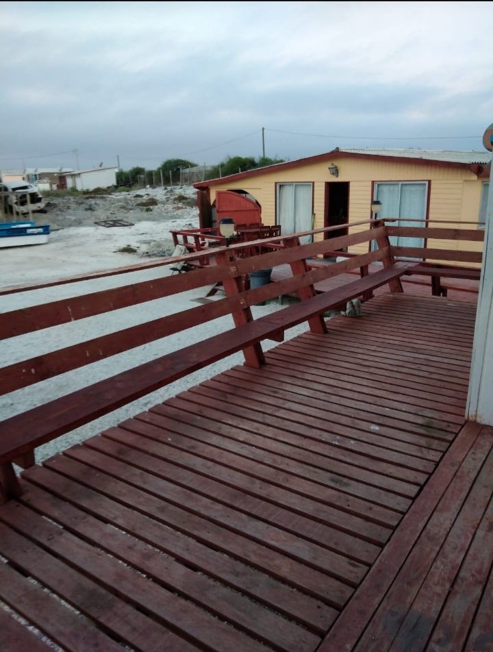 A wide wooden deck extends from the entrance, featuring a railing along its length. The deck leads to a yellow house with two large windows and a door. A small boat can be seen in the background, resting near the shore.