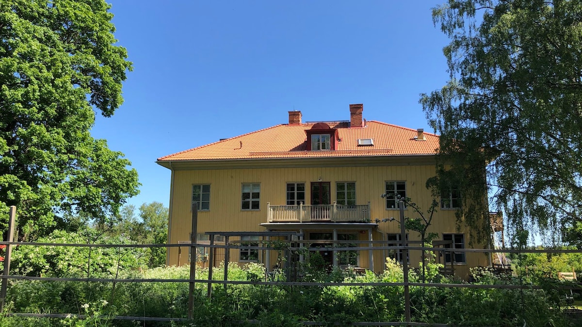 A historic yellow manor house is framed by lush green trees and vegetation, showcasing a red-tiled roof and multiple windows. A balcony features prominently on the second floor, and the surrounding landscape is filled with wildflowers and foliage.