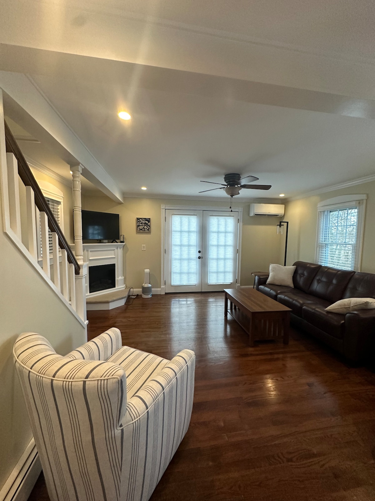 A welcoming living area is featured, showcasing a brown leather sofa and a striped armchair. Natural light fills the space through double French doors, while a wooden coffee table sits centrally. A ceiling fan and a wall-mounted television enhance the comfort of the room.
