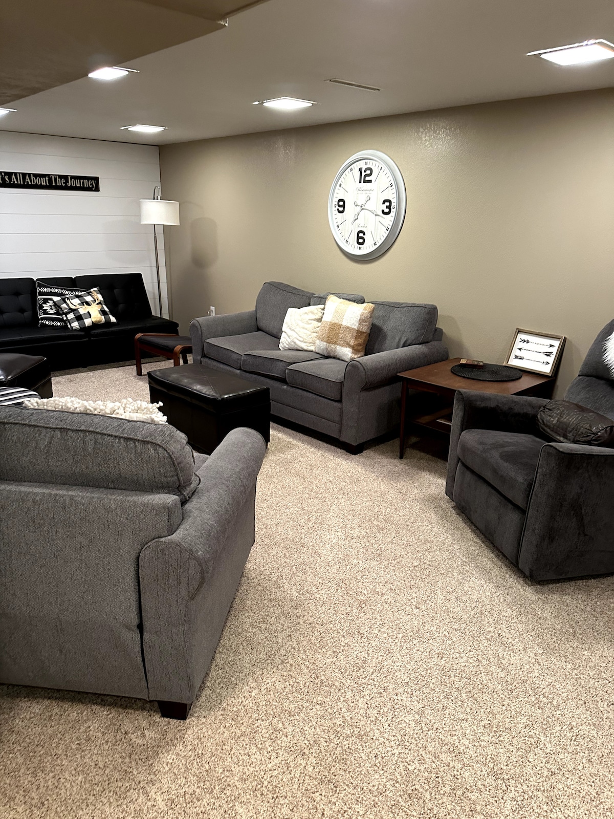 A cozy living area features grey upholstered sofas and armchairs arranged around a dark wooden coffee table. A large wall clock adds interest to the neutral-colored walls. Soft lighting enhances the inviting atmosphere, with a floor lamp providing additional illumination.