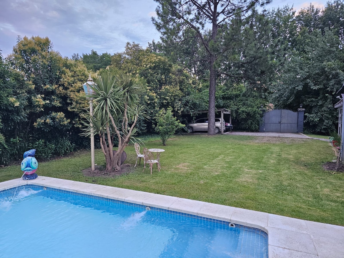 A clear view of a swimming pool is framed by lush greenery and a well-maintained lawn. Two small chairs and a table are placed near the pool, creating a serene outdoor space. Trees provide shade, enhancing the tranquil setting.
