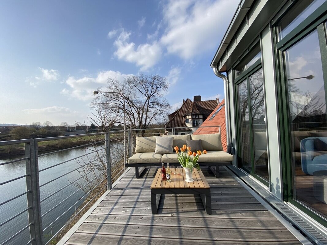 A spacious balcony is furnished with a comfortable seating area, featuring a sofa and a small table. Fresh flowers add a touch of color, while expansive windows provide views of the flowing river. The surrounding trees and rooftops create a serene outdoor setting.