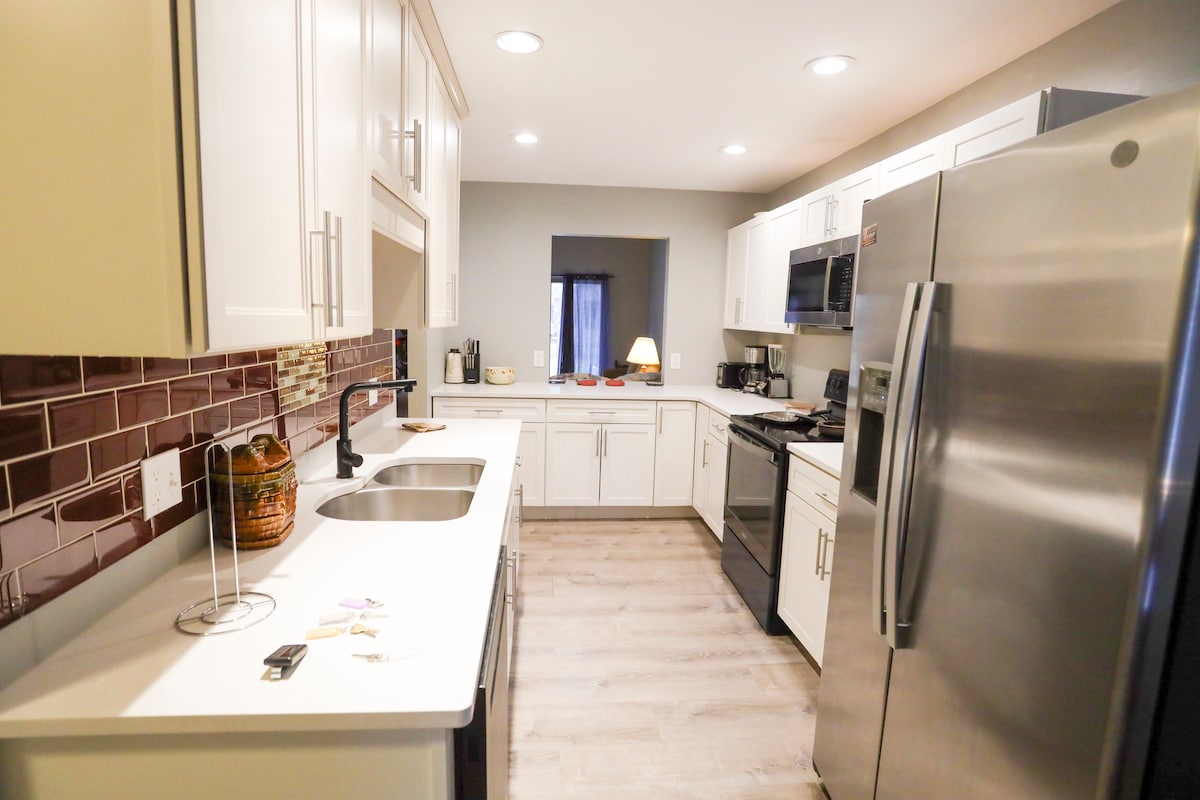 A modern kitchen is equipped with stainless steel appliances, including a refrigerator and stove. Sleek white cabinets and a light countertop create a clean look. A two-basin sink is positioned in front of a window, and textured brown tiles add warmth to the space.