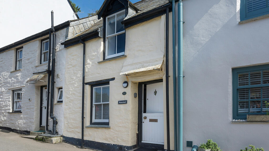 The exterior of the cottage is characterized by pale yellow stone walls and a classic black door framed by flanking windows. A pitched roof with slate tiles complements the quaint façade, inviting guests into a charming and historic space.