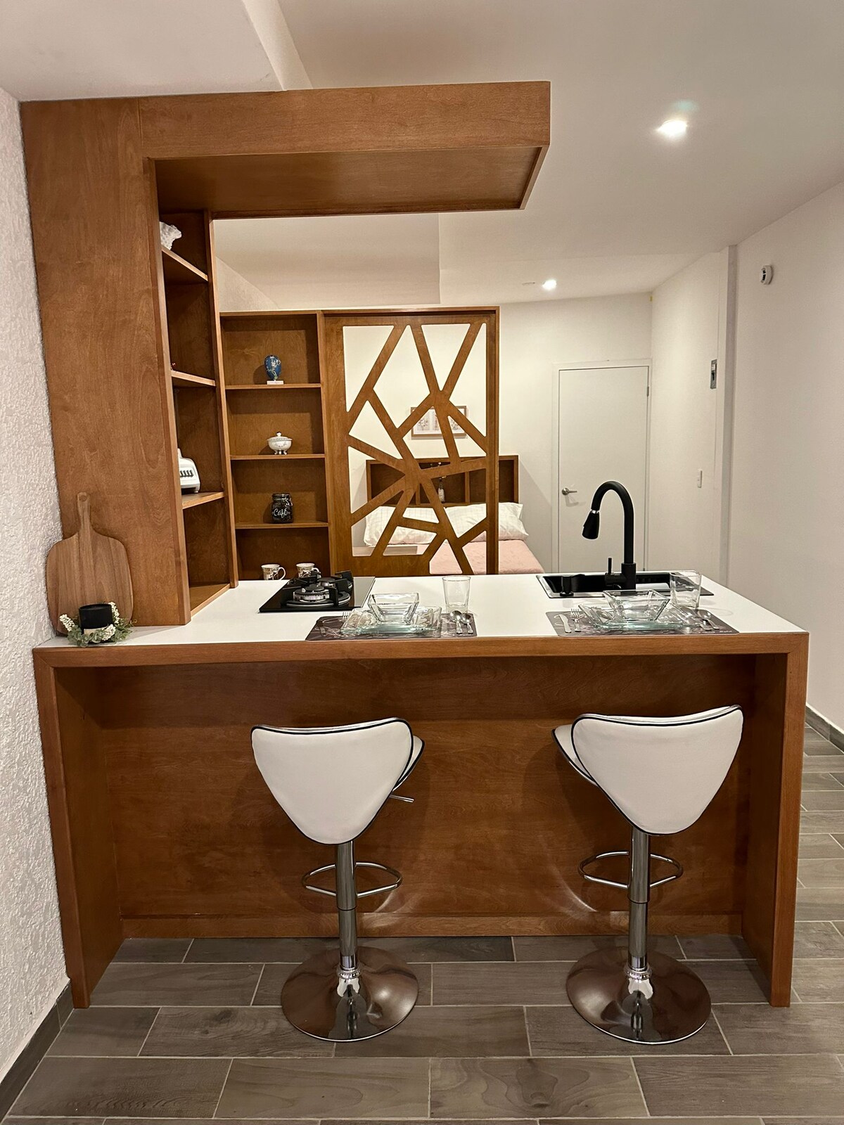 A modern kitchen area is showcased with a sleek countertop and two white bar stools. Wooden shelving is positioned above, displaying decorative items. A stylish black faucet is installed at the sink, blending functionality with contemporary design elements.
