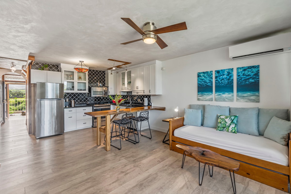 The open concept living area features a modern kitchen with stainless steel appliances and a dining table. A daybed with light blue cushions is positioned near the window, complemented by a decorative pillow. Large art pieces depicting ocean waves provide a calming visual.