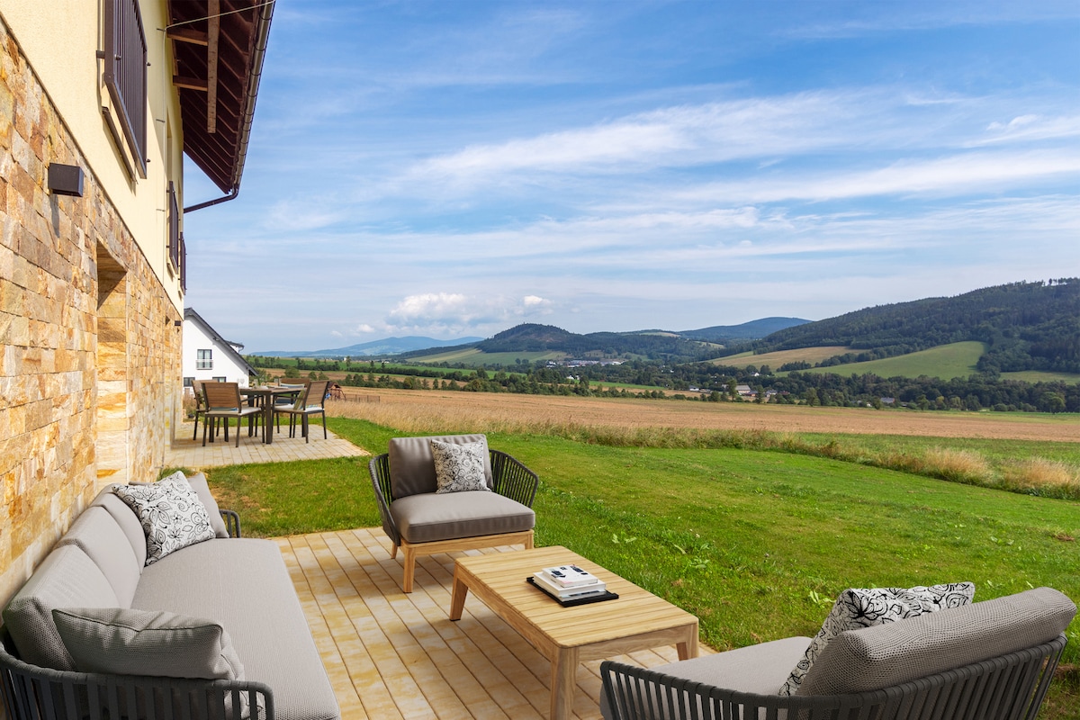 An outdoor terrace features comfortable seating with a coffee table, surrounded by expansive green fields and distant hills. A dining area is visible in the background, enhancing the serene landscape under a clear blue sky.