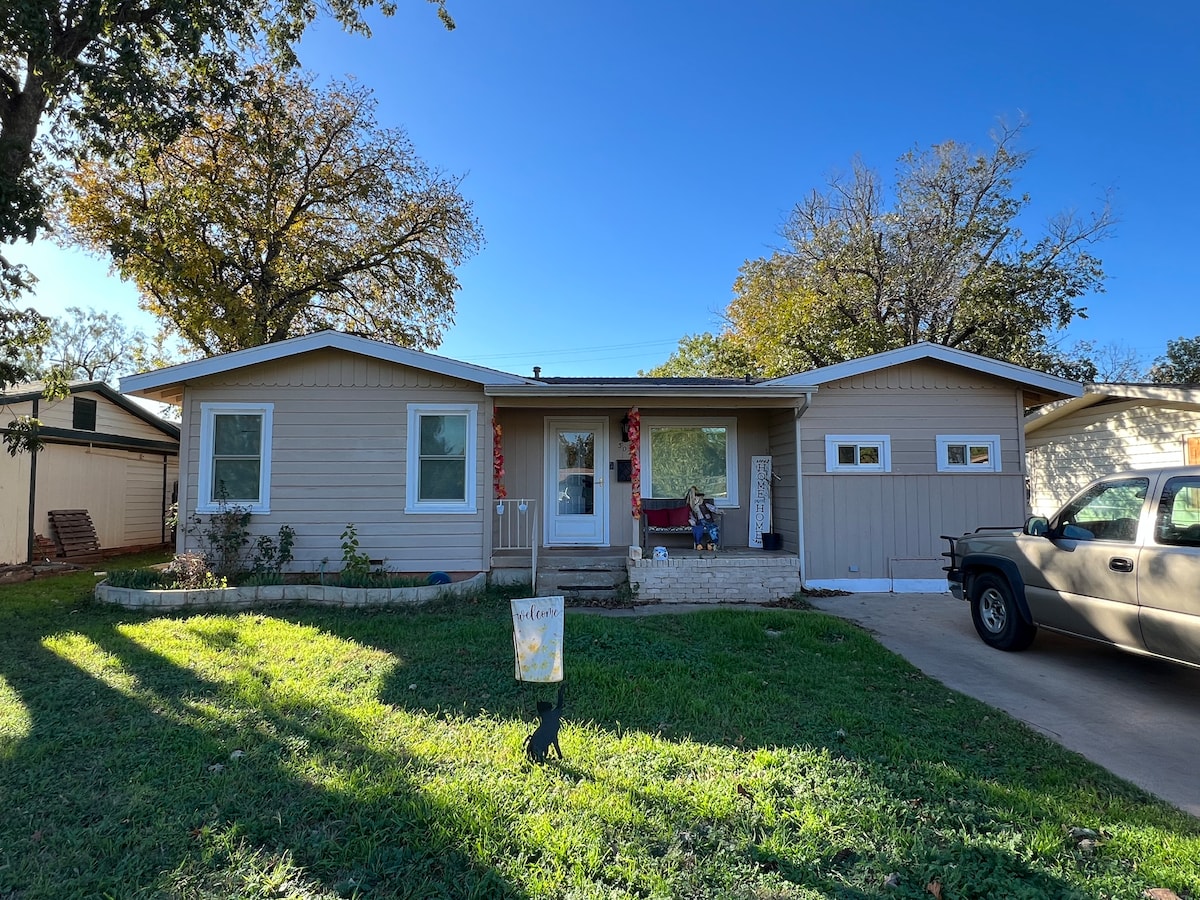 A welcoming single-story home is displayed with a light-colored exterior and a driveway leading to a garage. Neatly trimmed grass surrounds the front, which features a small garden and a porch accented by seasonal decorations. Trees provide shade nearby, enhancing the outdoor space.