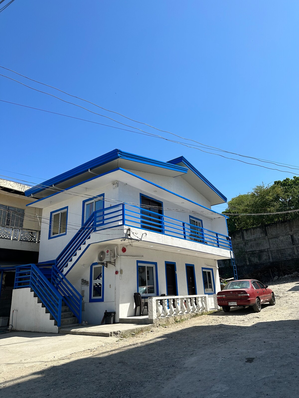The exterior of a two-story building is shown, featuring bright blue accents against a white facade. A staircase leads to the upper level, and large windows allow natural light to enter. The surrounding area includes a dirt pathway and a parked car.