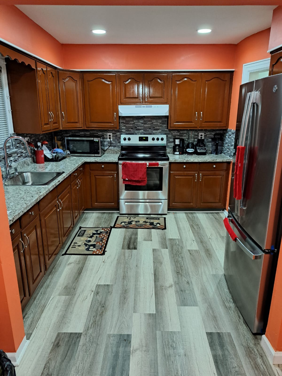 A well-appointed kitchen features wooden cabinets with a vibrant orange wall. Stainless steel appliances, including an oven and refrigerator, complement the granite countertops. A microwave and coffee maker are visible, along with two decorative rugs on the floor. Bright lighting enhances the spacious layout.