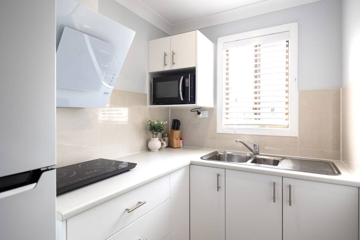 A modern kitchen space features white cabinetry and countertops. An induction cooktop is positioned near the sink, while a microwave is built into the cabinetry. Light streams in through the window adorned with wooden blinds, highlighting the neatly organized utensils on the counter.