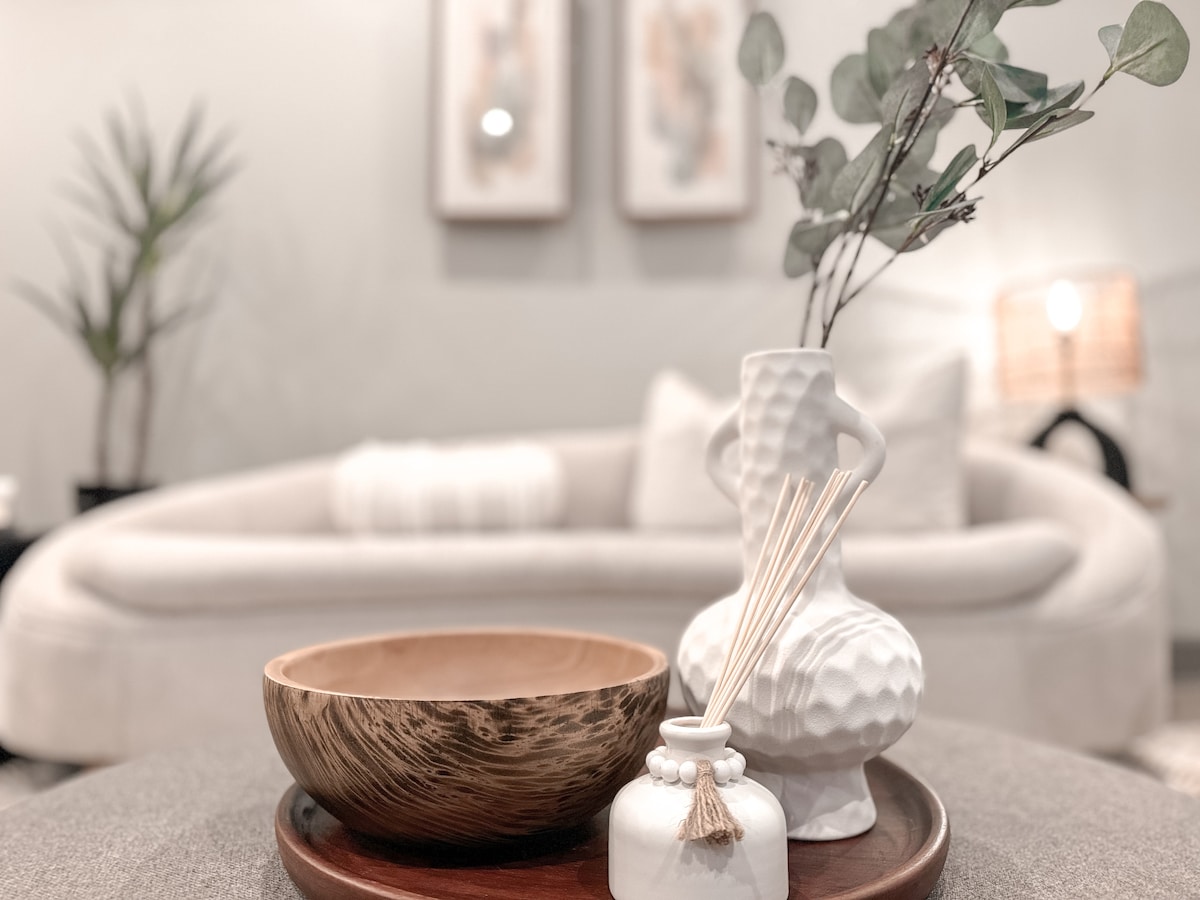 A decorative arrangement is showcased on a low table, featuring a wooden bowl and a textured white vase. Fresh greenery is displayed in the vase, while a small diffuser with reeds rests beside it. A stylish, curved sofa is visible in the background.