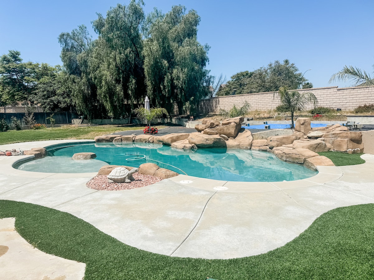 A landscaped outdoor area features a large, kidney-shaped pool surrounded by natural stone formations. Lush greenery and palm trees provide a tropical feel, while a smooth patio area offers ample space for relaxation and leisure activities. A separate pool is visible in the background.
