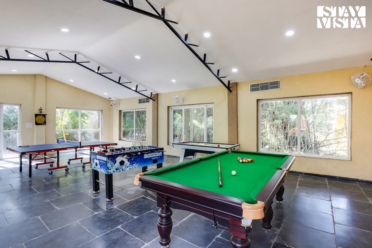 An entertainment area is showcased with various gaming tables, including a pool table and a foosball table. Spacious windows allow natural light to illuminate the room, revealing a serene view of the surrounding greenery. The floor is covered in dark stone tiles, enhancing the overall aesthetic.
