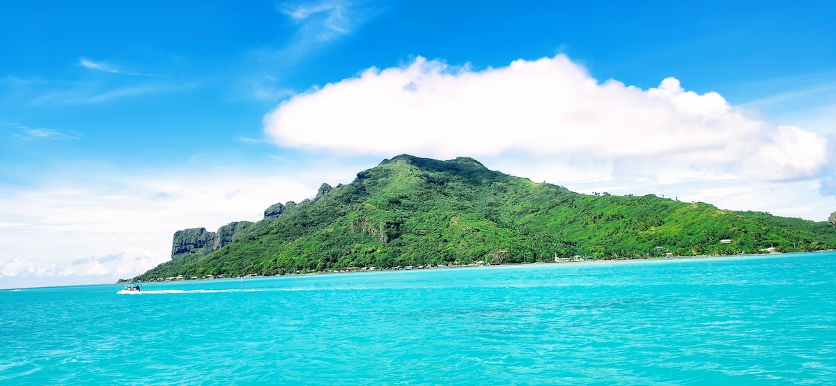 A vibrant blue ocean contrasts with the lush green landscape of the island in the distance. The backdrop features rocky cliffs interspersed with greenery, while a small boat glides through the water, showcasing the serene coastal environment.