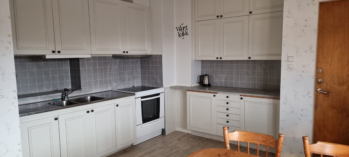 The kitchen features white cabinetry with a rustic design and grey tiled backsplash. A stainless steel kettle and an oven are visible, alongside a sink. A round wooden dining table with chairs is positioned, creating a functional and welcoming cooking space.
