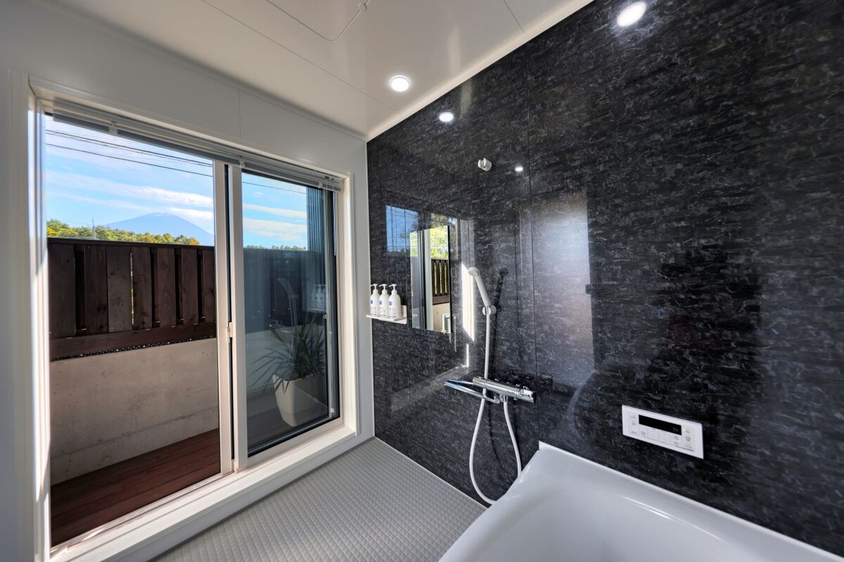 A modern bathroom features a spacious bathtub adjacent to a large window. Natural light streams in, illuminating sleek dark walls. A shower fixture is mounted on the wall, and elegant toiletries are arranged on a shelf nearby, providing a clean and functional space.