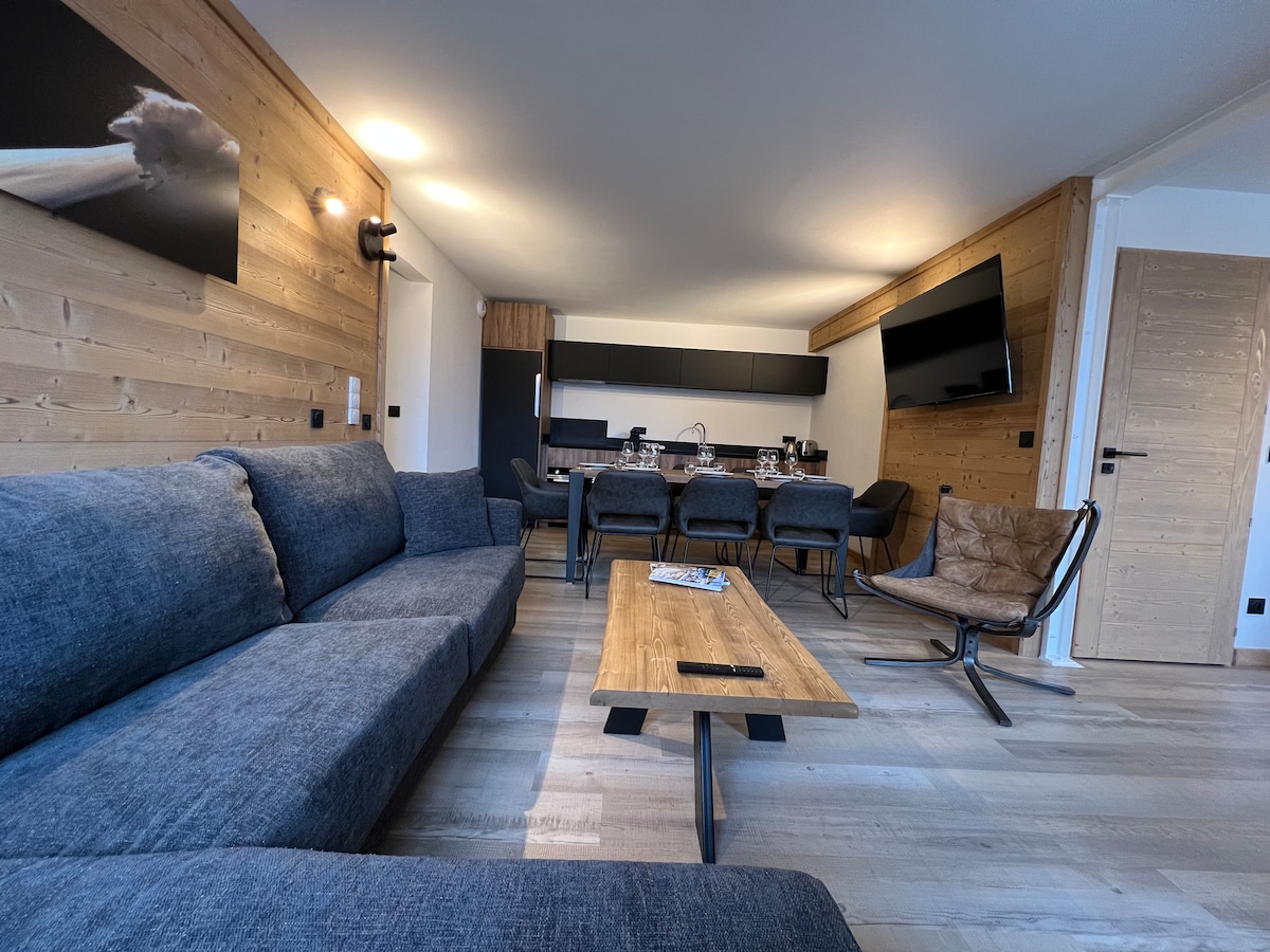 A spacious living area features a gray sectional sofa, a wooden coffee table, and a dining space with dark chairs. Wall-mounted artwork and a flat-screen television are visible, contributing to a modern and comfortable ambiance.