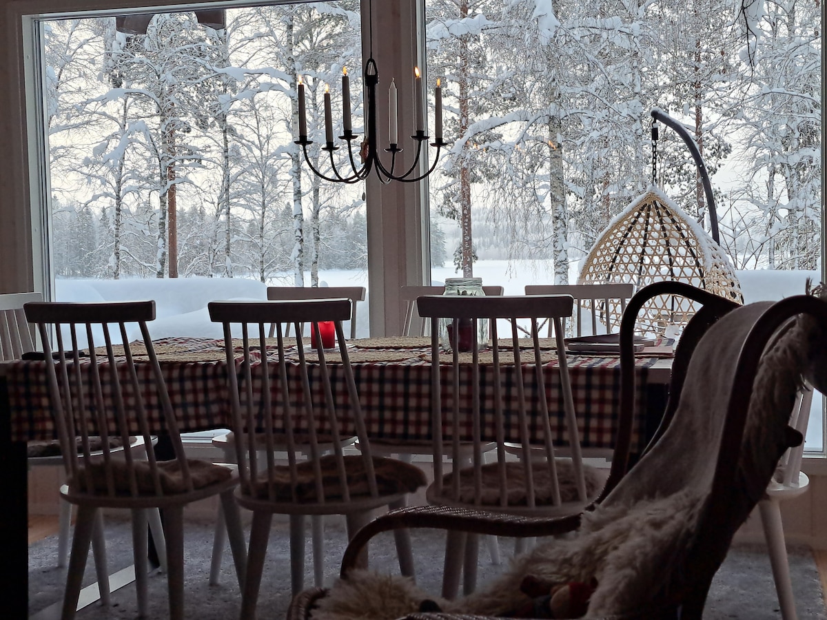 A spacious dining area is featured, with a long table set against large windows showcasing a snowy landscape. A chandelier hangs above the table, while a cozy chair sits nearby, contributing to the warm interior ambiance.