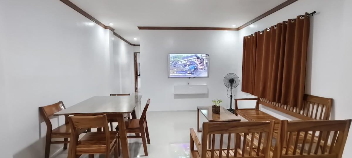 A bright living and dining area features wooden furniture, including a dining table with four chairs and a comfortable seating arrangement. A wall-mounted TV is positioned across from the seating, and a fan is nearby. Natural light filters through brown curtains at the window.