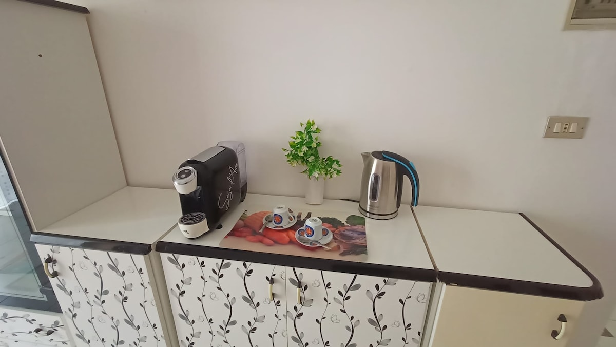 A compact kitchenette is presented, featuring a coffee machine and electric kettle on a counter. A small decorative plant adds a touch of greenery. Nearby, two cups are placed on a colorful mat, contributing to the overall functionality of the space.