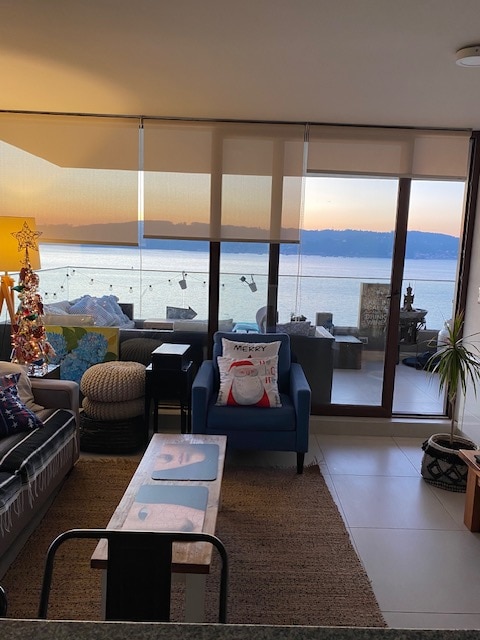 A cozy living area features a large window showcasing a panoramic view of the bay at sunset. Comfortable seating is arranged around a wooden coffee table. Decorative items, including a small Christmas tree and a pillow with a festive design, add unique touches to the space.