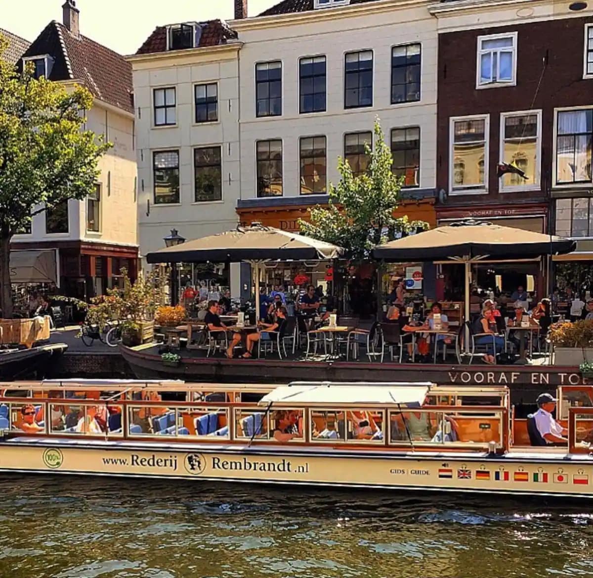 A lively riverside scene is depicted, featuring a boat that navigates the waterway. Outdoor seating at nearby cafes is visible, with patrons enjoying their time. Trees line the bank, enhancing the vibrant atmosphere of the location, reflecting the charm of the surrounding architecture.