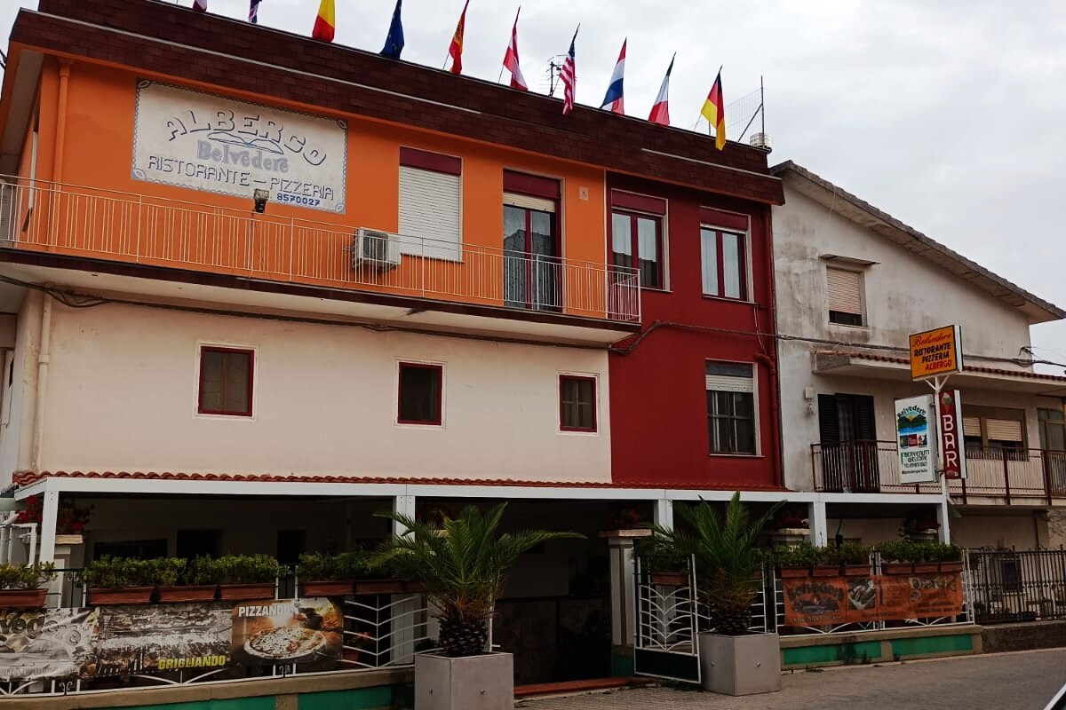 The building features a two-story facade with a mix of colors, including red and white. Flags of various nations are displayed prominently along the top. The ground level highlights a restaurant and pizza signage, complemented by potted plants along the entrance.