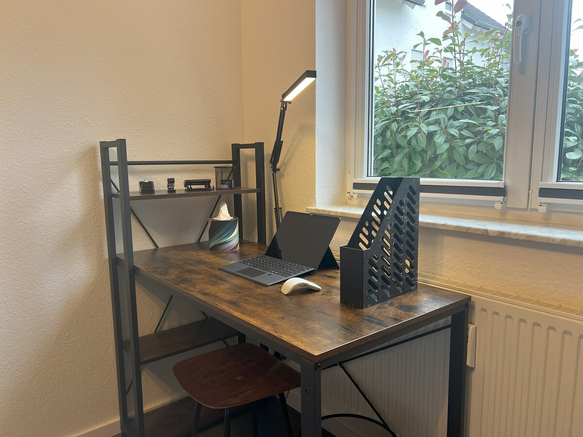 A work desk is positioned by the window, featuring a laptop, a mouse, and a lamp. The desk's design includes an open shelf for storage. Light filters in through the window, illuminating the workspace, which overlooks greenery outside.