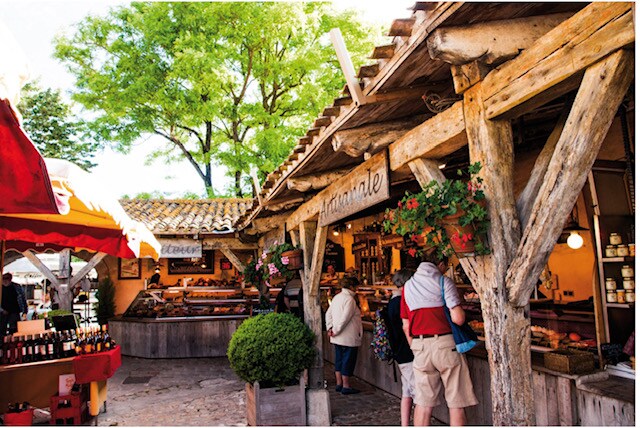 A charming market scene features rustic wooden stalls adorned with vibrant awnings. Fresh produce, local delicacies, and handmade goods are displayed prominently. Patrons are engaged in shopping, surrounded by greenery and a bustling atmosphere. The structure exhibits wood beams and potted plants adding to the overall appeal.