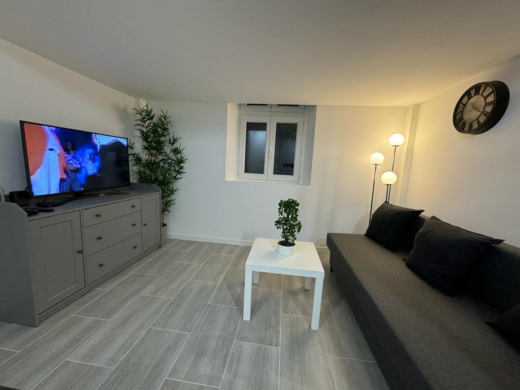 A cozy living area features a dark gray sofa adorned with black cushions. A small white table is placed in front of the sofa. A flat-screen television is mounted on the wall opposite, alongside a decorative plant. Light is provided by a tall floor lamp.