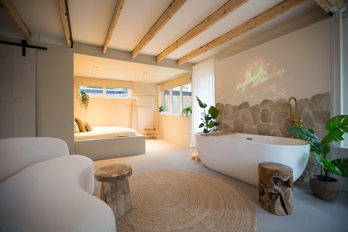 A bright and open living space features a spacious bed, a standalone bathtub, and a cozy seating area. Natural materials are used, with wooden beams overhead and stone accents on the wall. Soft lighting and greenery enhance the serene ambiance.