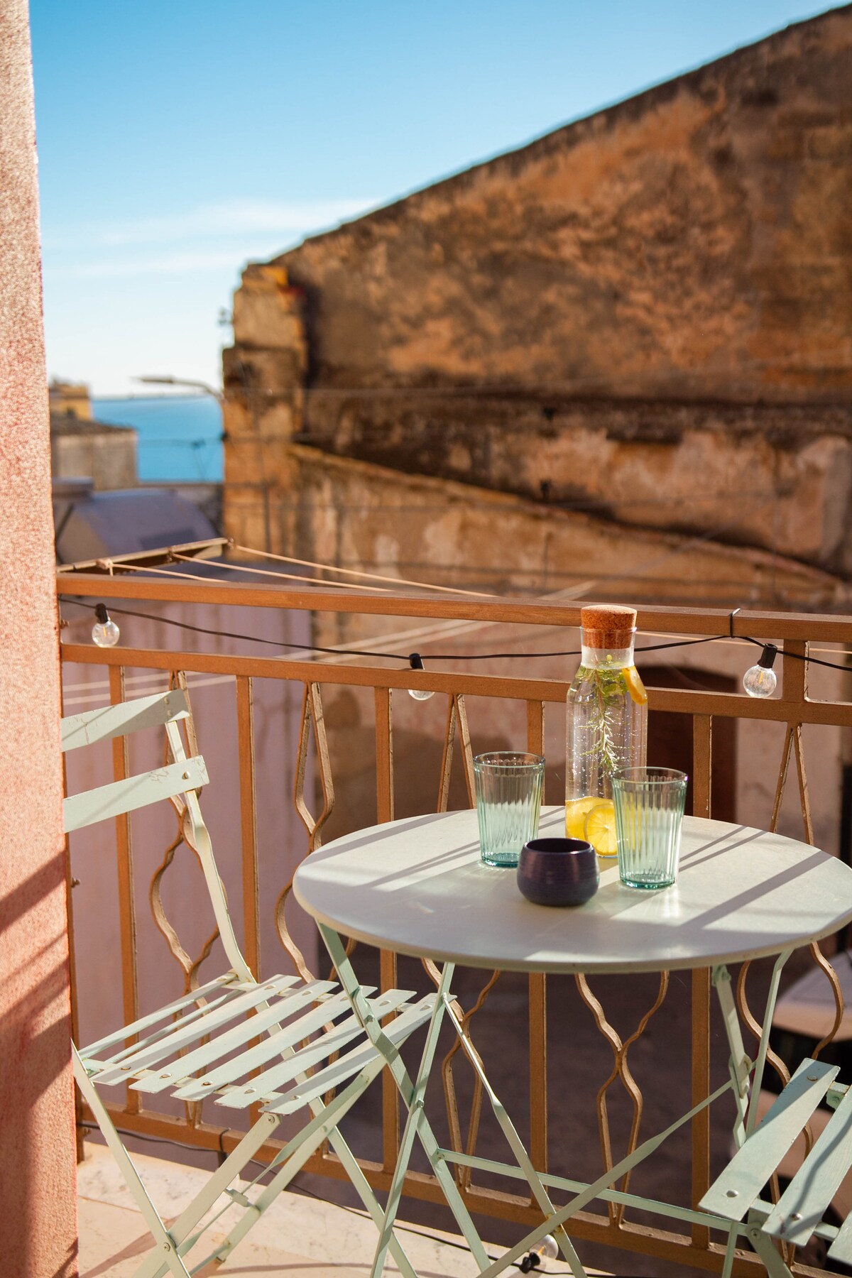 A small balcony is furnished with a round table and two mint-colored metal chairs. A glass water bottle and two drinking glasses are placed on the table. The background features a glimpse of blue sky and rooftops, enhancing the peaceful surroundings.