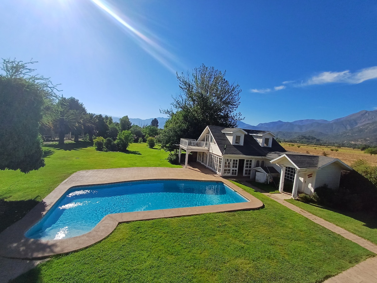 A large outdoor area features a clear blue pool surrounded by a manicured lawn. The poolside area is accented by a covered patio with seating, and a charming white house is situated in the background, framed by trees and distant mountains.