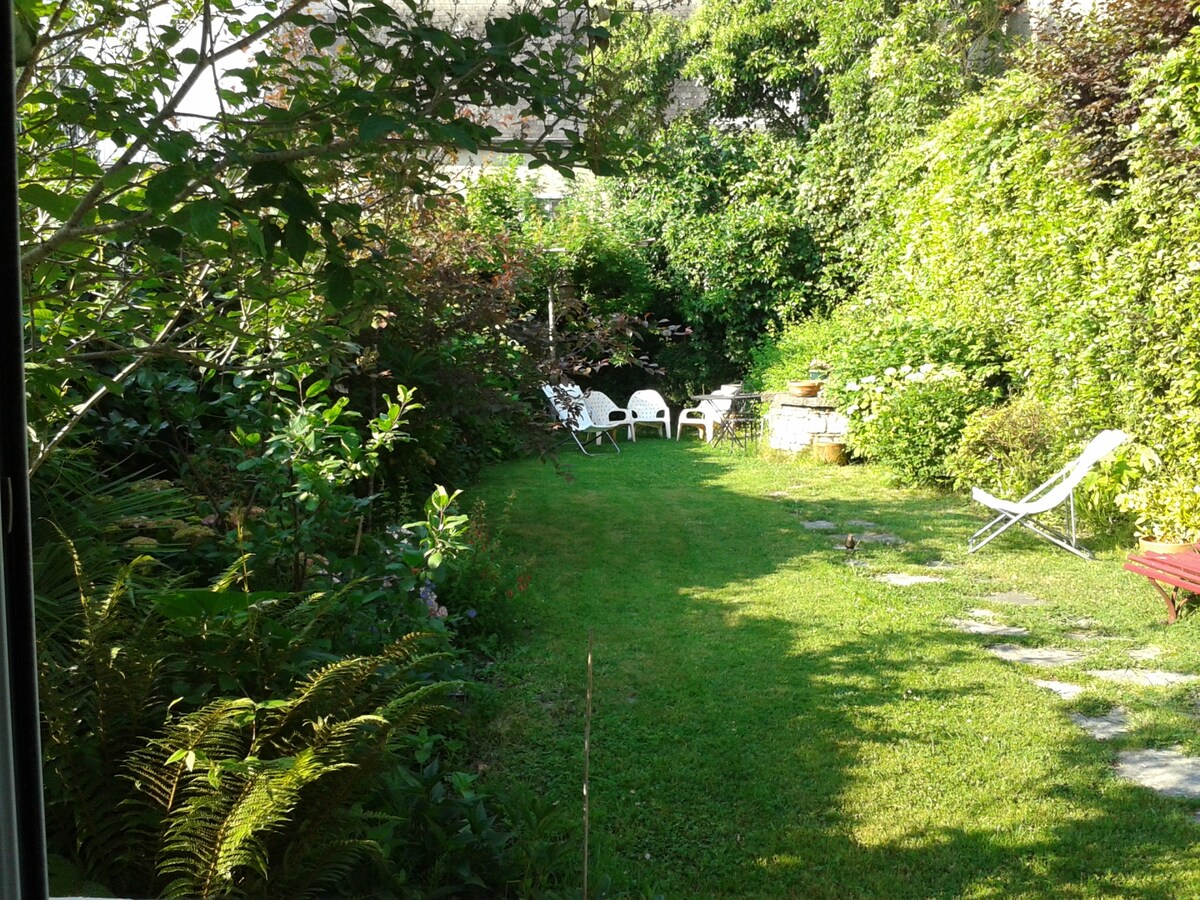 A lush garden area is seen, featuring well-maintained grass surrounded by diverse greenery. Several white chairs are positioned for seating, with a lounge chair visible. A stone path leads through the garden, inviting relaxation amid the vibrant plant life.