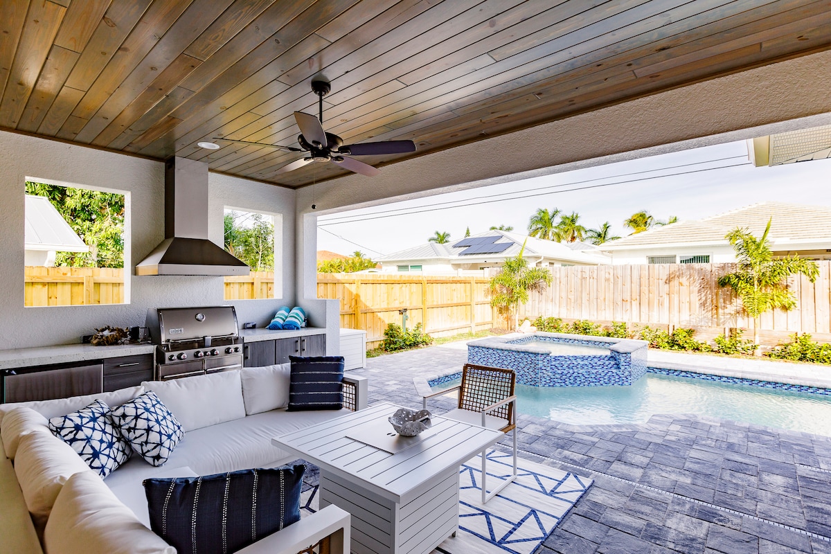 An outdoor living space features a covered seating area with a sectional sofa adorned with decorative pillows. A modern grill is positioned nearby, and a pool with a unique design is visible in the background, surrounded by lush greenery and a wooden fence.
